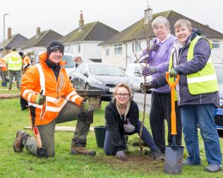 A man kneeling in high vis with thumbs up with three smiling women next to a recently-planted tree.
