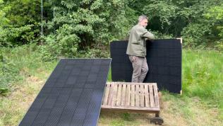 A man in a leafy green clearing carries a solar panel towards an empty space in a structure on the ground that is already holding up a second solar panel at an angle.