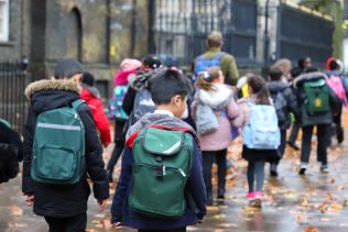 A group of primary school age children walk along a pavement with their teacher.