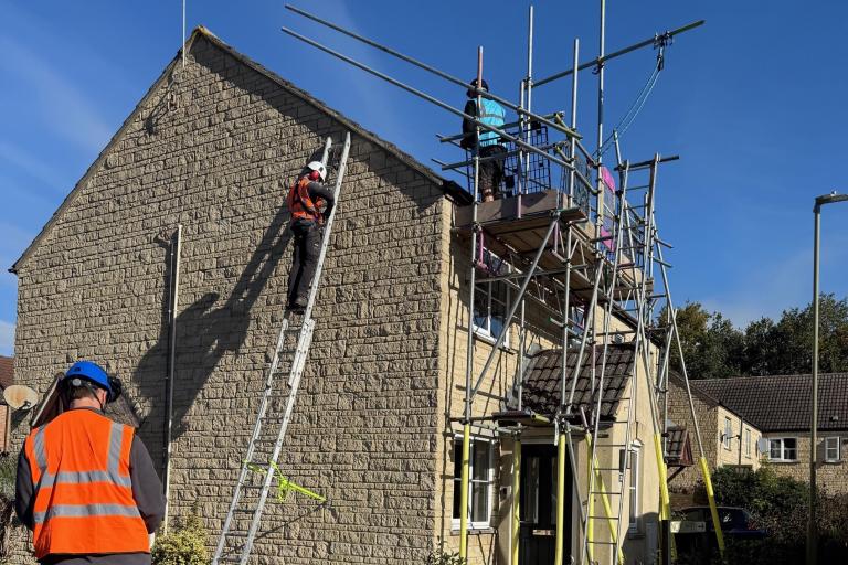 A pale brick house with scaffolding at the front, and a man in an orange hi-vis and hard hat on a ladder at the side, climbing up to the roof.