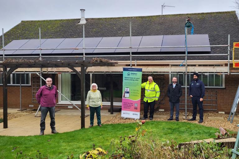 Five smiling people stand in front of a house and its newly-installed solar panels, scaffolding still in place.