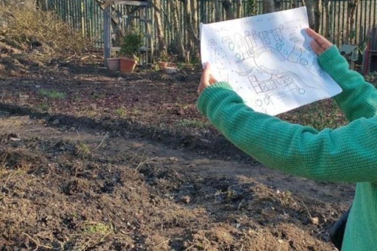 An empty plot of land with someone holding up some plans for it.