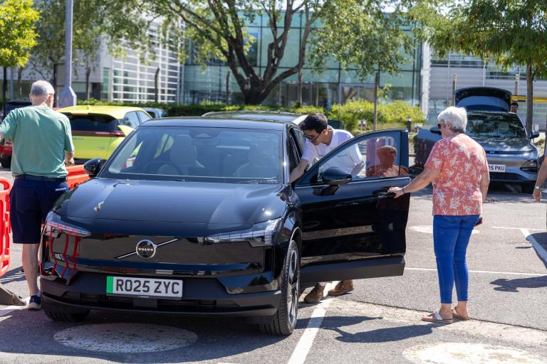 A black EV car sits in a car park with it's doors open on a sunny day as three people have a look inside it.