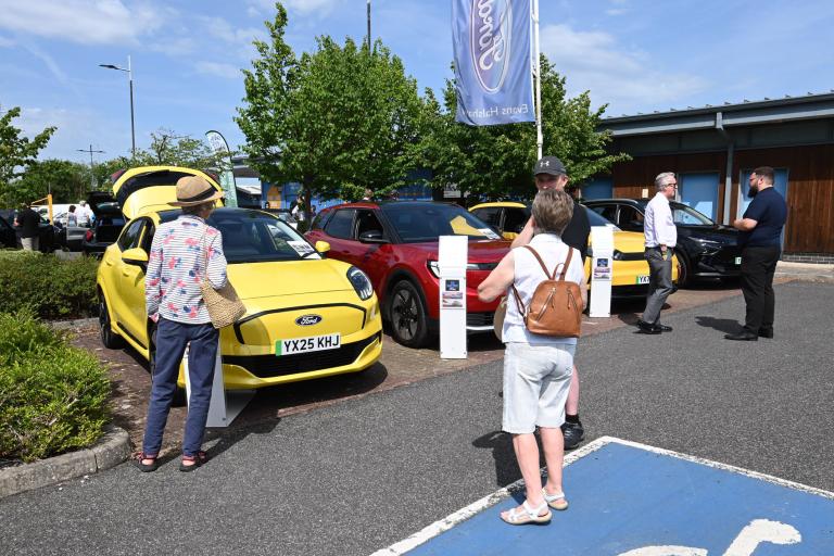 Four EV cars sit in a row parked in a car park on a sunny day as five people take a look at them.