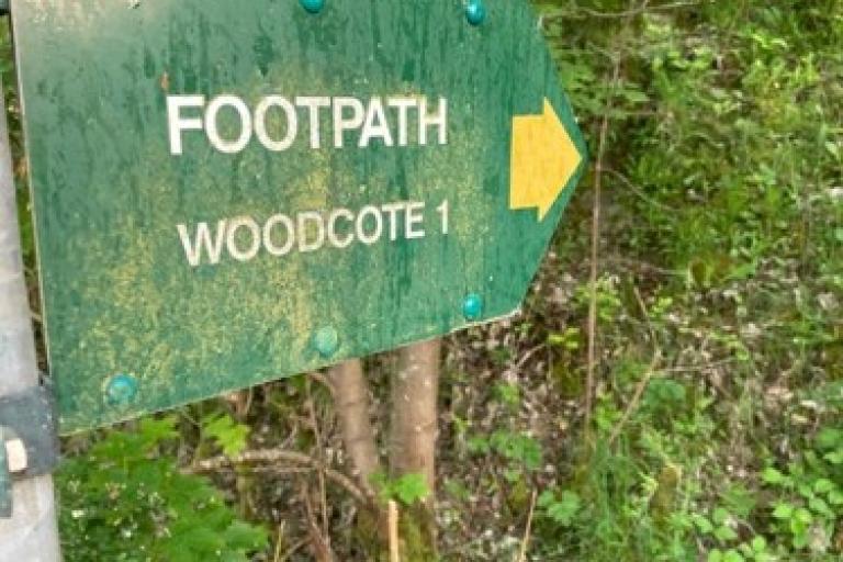 A footpath sign reading 'woodcote 1' sits amongst some greenery.