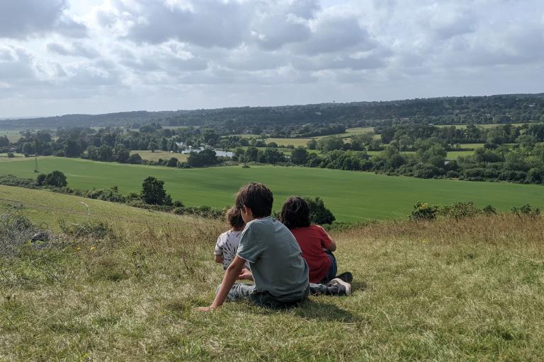 Three children sit on the grass with rolling hills in the background.